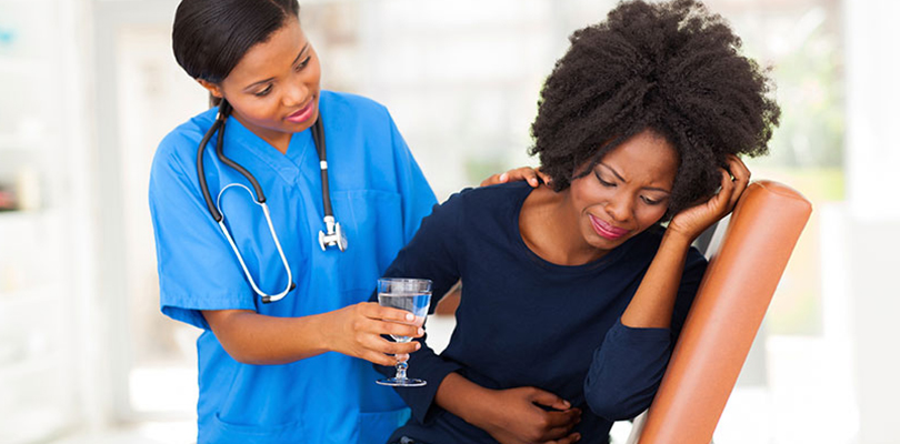 Woman is holding her stomach because of abdominal pain and a doctor is beside her holding a glass of water