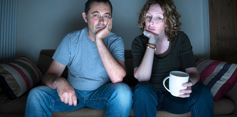 A couple is sitting on a couch watching TV looking tired