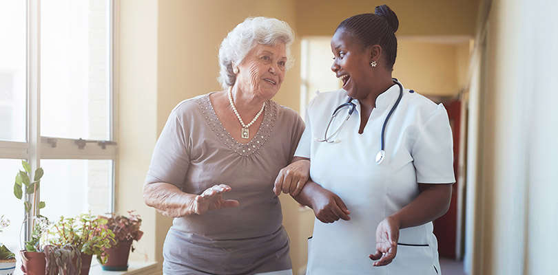 A caregiver and an elderly woman are walking and talking in an assisted living home